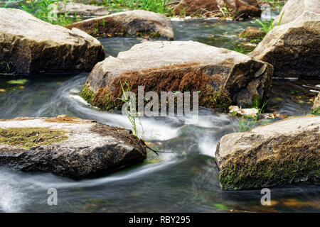 Detailansicht des fließenden Wasser eines kleinen Flusses, fließt das Wasser zwischen großen Steinen, teilweise mit Moos und Gras bewachsen, Langzeitbelichtung Stockfoto