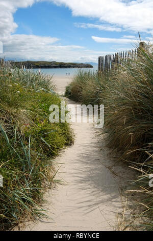 Ein sandiger Pfad mit Gras Schilf auf jeder Seite und einem Holzzaun auf der rechten Seite führt zu einer wunderschönen Bucht mit ruhigem blauen Meer und eine Insel in der middl Stockfoto