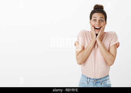 Froh und glücklich Junges attraktives Mädchen mit unordentlichen Bun in Pink stylische T-Shirt überrascht, Hände auf die Wangen fröhlich lächelnd und optimistisch Anhörung erstaunlich gute Nachrichten über graue Wand Stockfoto