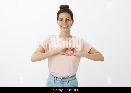 Unterstützende und fürsorgliche Freundin im stilvollen t-shirt mit unordentlichen hairbun zeigen Herz Geste in der Nähe der Brust als bekennende in Liebe und Bewunderung lächelte breit mit sanften Grinsen an Kamera Stockfoto