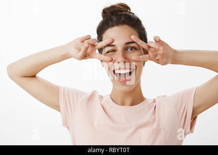 Lassen Sie uns diese Party Rock. Aktive und glückliche junge optimistisch attraktive Frau mit bun Frisur in t-shirt, mit Freude an der Kamera winking, Frieden oder Sieg Geste über Augen und breit grinsend Stockfoto