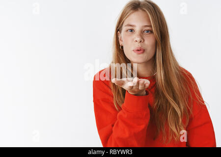 Senden von warmen Kuss über die Linse der Kamera. Charmantes Angebot Freundin mit roten Haaren, blauen Augen und Sommersprossen falten Lippen zu senden Luft mwah, holding Palm in der Nähe von Gesicht, Schweiß und Pflege über graue Wand Stockfoto