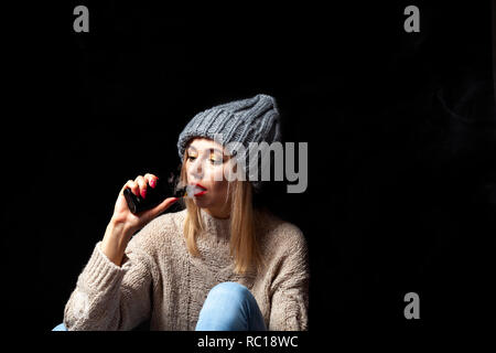 Blondine in einen gestrickten Pullover und Hut hält eine elektronische Zigarette vape in ihrer Hand mit einer Maniküre und atmet Rauch aus ihrem Mund, die Lippen sind Schmerzen Stockfoto