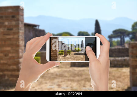 Ein Tourist ist ein Foto von einer Straße ohne Menschen mit den Ruinen der Häuser in Pompeji, mit dem Vesuv im Hintergrund auf einem Mobiltelefon Stockfoto