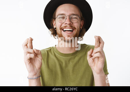 Taille - geschossen von optimistisch sorglos und hübscher junger bärtiger Kerl in der Brille und Hut laut lachend mit freudiger Ausdruck als drückt die Daumen für gutes Glück, die Traum über graue Wand kommen Stockfoto