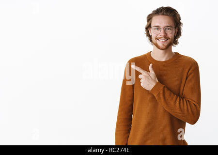 Gut aussehender bärtiger Kerl mit gewelltes Haar in Pullover und Gläser freudig lächelnd als Empfehlung statt nach links, einladende Freund schnappen Biss und sprechen gerne über grauer Hintergrund posiert Stockfoto