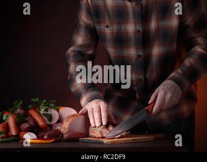 Mann in einem brown Plaid Shirt schneidet verschiedene Wurst und geräuchertes Fleisch auf einem alten Holztisch. Fleisch, Gewürze und Kräuter auf einem Küchentisch. Kopieren Sie Platz. Stockfoto