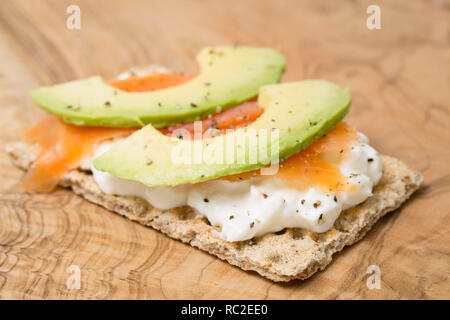 Ein Roggen Brot Cracker mit Fett frei Cottage Käse, in Scheiben geschnittenen Avocado pear und bewirtschafteten, geräucherter Lachs Scheiben mit schwarzer Pfeffer. Auf eine Olive Holz hacken Stockfoto