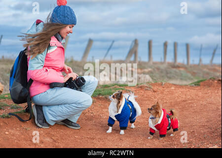 Schöne junge Reisende Frau in Wolle hat Trainingsanzug und Rucksack mit Kamera beobachten an Ihrem kleinen gekleidet Chihuahua hunde während Outdoor hi lächelnd Stockfoto