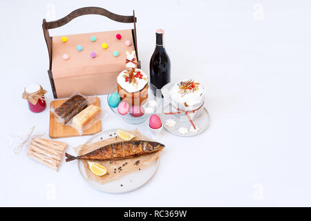 Ostern Kuchen, bunte Eier, geräucherten Fisch und eine Flasche Rotwein. Orthodoxe süßes Brot, kulich und anderen traditionellen Nahrungsmitteln auf hellen Hintergrund. Geschenke für Urlaub in Holz- Korb. Copyspace für Text Stockfoto