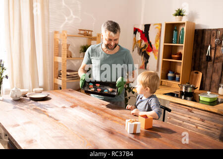 Lächelnd schöne bärtige Vater holding Pan und handgefertigten Sugar Cookies zu kleinen Sohn auf den Küchentisch Stockfoto