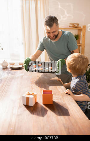 Fröhliche bärtigen Vater im Ofen Mitt unter Pan mit Valentines cookies aus dem Ofen und am Tisch, während Geschenk gemeinsam mit Sohn Stockfoto