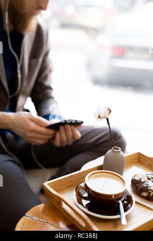 In der Nähe von Holz- Fach mit morgendlichen Cappuccino und Schokolade Donut, hipster Mann im Hintergrund smartphone Stockfoto