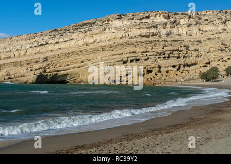 Strand mit Sand und Wellen und einer Klippe mit alten Höhlen im Hintergrund vor blauem Himmel, Bilder von Matala im Süden der Insel Kreta Griechenland. Stockfoto