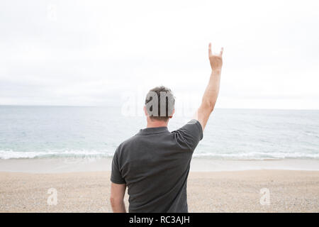 Eine Ansicht von hinten von einem starken jungen Mann hob seinen Arm in die Luft und ein Teufel horn Geste, während mit Blick auf den Ozean in ein Symbol der Hoffnung und Defiance Stockfoto