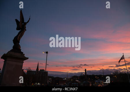 Kidderminster, Großbritannien. 13. Januar, 2019. UK Wetter: die Sonne in Kidderminster nach einem ungewöhnlich warmen Tag mit Temperaturen um 10 Grad Celsius. Die Skulptur der Engel des Friedens sehen Sie hier die Hintergrundbeleuchtung durch die bunten Sonnenuntergang außerhalb der Kirche St. Mary, Kidderminster. Quelle: Lee Hudson/Alamy leben Nachrichten Stockfoto