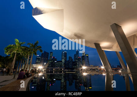 Lit ArtScience Museum gesehen von unten und Reflexionen der Wolkenkratzer im Geschäftsviertel in Singapur in der Abenddämmerung. Mehrere Personen sitzen und stehen neben dem Pool. Stockfoto