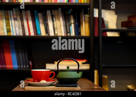 Chinesischer grüner Tee pot und rot Tasse Kräutertee Auf der rauhen Holztisch mit Bücherregalen auf dem Hintergrund. Erholsame Zeit von Reisen und Stockfoto