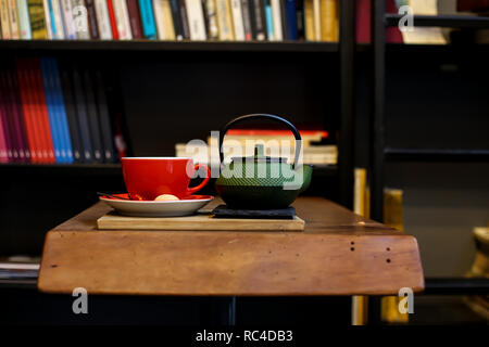 Chinesischer grüner Tee pot und rot Tasse Kräutertee Auf der rauhen Holztisch mit Bücherregalen auf dem Hintergrund. Erholsame Zeit von Reisen und Stockfoto