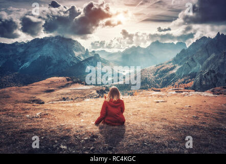 Junge Frau sitzt auf dem Feld gegen die majestätischen Berge bei Sonnenuntergang im Herbst in den Dolomiten, Italien. Landschaft mit Mädchen, bewölkter Himmel, orange Gr Stockfoto