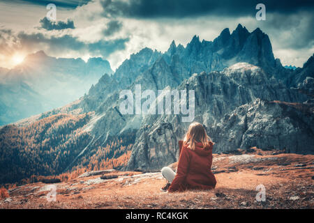 Junge Frau sitzt auf dem Hügel gegen die majestätischen Berge bei Sonnenuntergang im Herbst in den Dolomiten, Italien. Landschaft mit Mädchen, bewölkter Himmel, orange gra Stockfoto