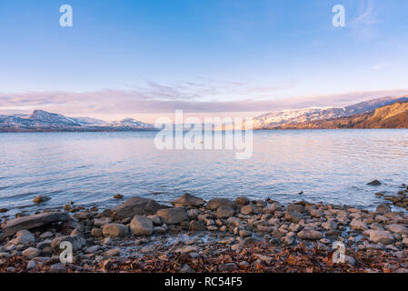 Felsige Strand mit Blick auf den See und die schneebedeckten Berge bei Sonnenuntergang Stockfoto