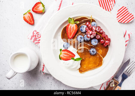 Pfannkuchen in der Form eines Herzens mit Beeren, Obst und Quark Sahne in eine weiße Platte, Ansicht von oben. Valentinstag Konzept. Stockfoto