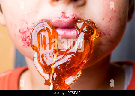 Boy saugt Lollipop, Baby isst - rot-orange Chupa Chups, Mund, rosa Lippen, Süßigkeiten in der Hand, das Gesicht halb Junge Stockfoto