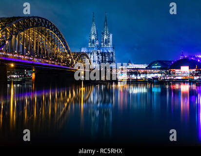 Die Hohenzollernbrücke (l), der Köllner Dom (m) und der Musical Dome (r) spiegeln sich bei blauen Stunde in Regenwasser in einer Mauer. Stockfoto