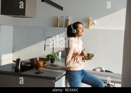 Fröhlicher junger Mädchen Musik hören mit Kopfhörern in der Küche zu Hause, Essen Salat aus einer Schüssel Stockfoto