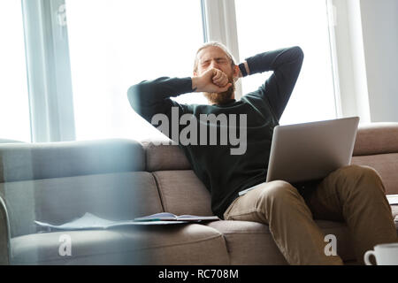 Bild von einem müden Jungen bärtigen Mann sitzt mit Laptop zu Hause. Stockfoto