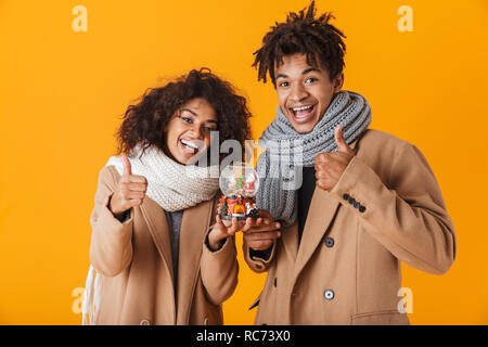 Gerne afrikanische Paar tragen Winter Kleidung über Gelb Hintergrund isoliert, die hielten Weihnachten Schneekugel, Daumen hoch Stockfoto