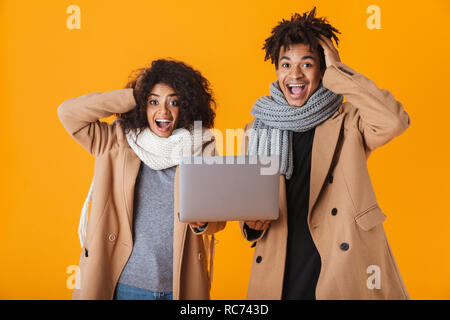 Gerne afrikanische Paar tragen Winter Kleidung über Gelb Hintergrund isoliert, die hielten Laptop Stockfoto