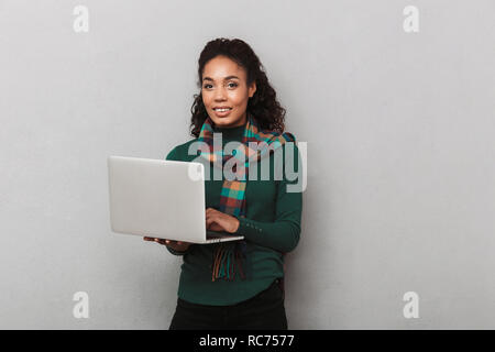 Fröhlich afrikanische Frau mit Kopftuch stehend über grauen Hintergrund isoliert, Laptop Stockfoto