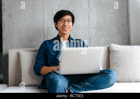 Lächelnden jungen asiatischer Mann auf einer Couch mit Laptop zu Hause sitzen Stockfoto