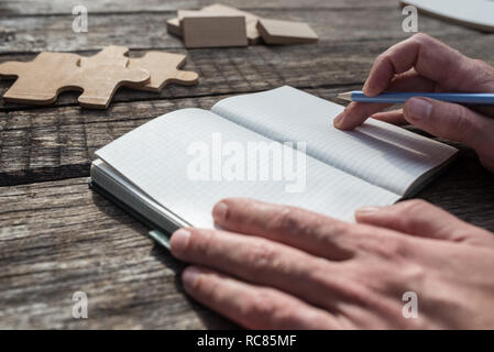 Nahaufnahme eines Studenten Brainstorming schreiben Ideen in seinem Notizblock mit Holz Puzzleteile und Stifte auf seine rustikalen hölzernen Schreibtisch platziert. Stockfoto