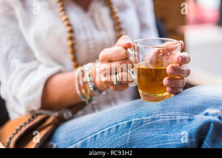 Nahaufnahme von Frau Hände unter chamomil camomilla Glas zum Aufwärmen und Entspannen - lässige Jeans und Pfingstmontag eshirt - Outdoor Freizeitaktivitäten Aktivität - Trinken heilen Stockfoto