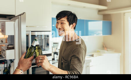 Lächelnd asiatischer Mann zu Hause stehend Toasten eine Flasche Bier mit seinem Freund. Freunde eine Party zu Hause mit Bierflaschen. Stockfoto