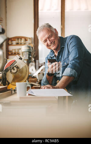 Senior Zimmermann in seiner Werkstatt mit Handy lächelnd. Reifer Mann in seiner Schreinerei lesen SMS-Nachricht auf sein Smartphone. Stockfoto