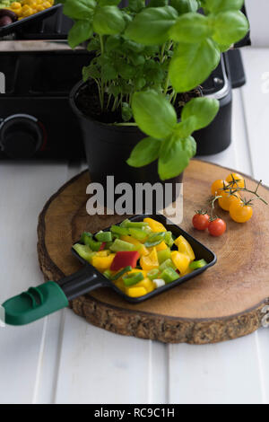 Vegan Raclette, raclette Pan mit fein gehackten Gemüse auf einem Holzbrett neben Tomaten und einen Topf Basilikum Stockfoto