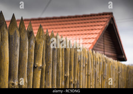 Ein Zaun aus spitzen Holzpfählen vor dem Hintergrund der ein Holzhaus mit einem roten Ziegeldach. Stockfoto