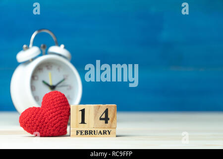 Valentines Tag Konzept. hand Garn machen Rotes Herz neben Holzblock Kalender auf Valentines Datum 14. Februar vor der weißen Wecker auf Holz gesetzt Stockfoto