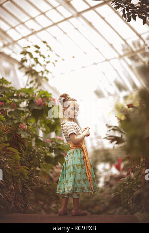 Kleines Mädchen in einem bunten Kleid von Pflanzen in einem Gewächshaus umgeben Stockfoto