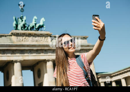 Junge schöne positive Mädchen macht selfie vor dem Hintergrund des Brandenburger Tor in Berlin in Deutschland oder nimmt Bilder von Sehenswürdigkeiten. Stockfoto