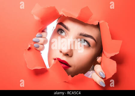 Das Gesicht der jungen schönen Mädchen mit einem hellen Make-up und mit dicken roten Lippen Einblicke in ein Loch in der orange Papier Stockfoto