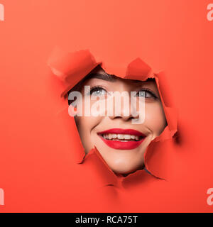 Das Gesicht der jungen schönen Mädchen mit einem hellen Make-up und mit dicken roten Lippen Einblicke in ein Loch in der orange Papier Stockfoto