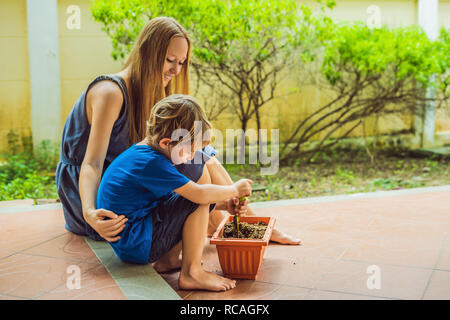 Schöne junge Frau und ihres süßen Sohn einpflanzen Sämlinge im Bett in den heimischen Garten im Sommer Tag. Garten Werkzeug, Handschuhe und Gießkanne im Freien. Gartenbau Aktivität mit Kind und Familie Stockfoto