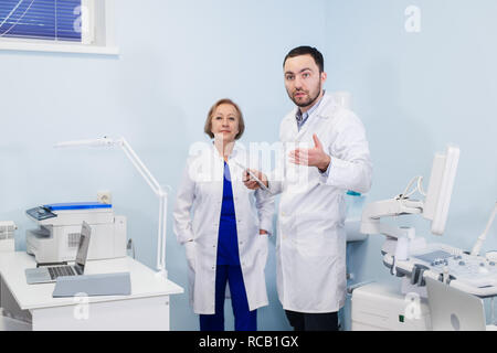 Leitender Arzt im Gespräch mit jungen Mann Assistant in der gynäkologischen Office mit Stuhl und Lampe auf dem Hintergrund Stockfoto