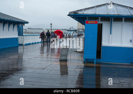 Menschen zu Fuß bei Regen hinter einer Herrentoilette auf Llandudno Pier, Conwy, Wales, Großbritannien Stockfoto