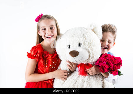 Hübsches kleines Mädchen und Jungen feiern Valentinstag und halten rote Rosen Schaufel und der weiße Bär Geschenk auf dem weißen Hintergrund mit roten Herzen Stockfoto